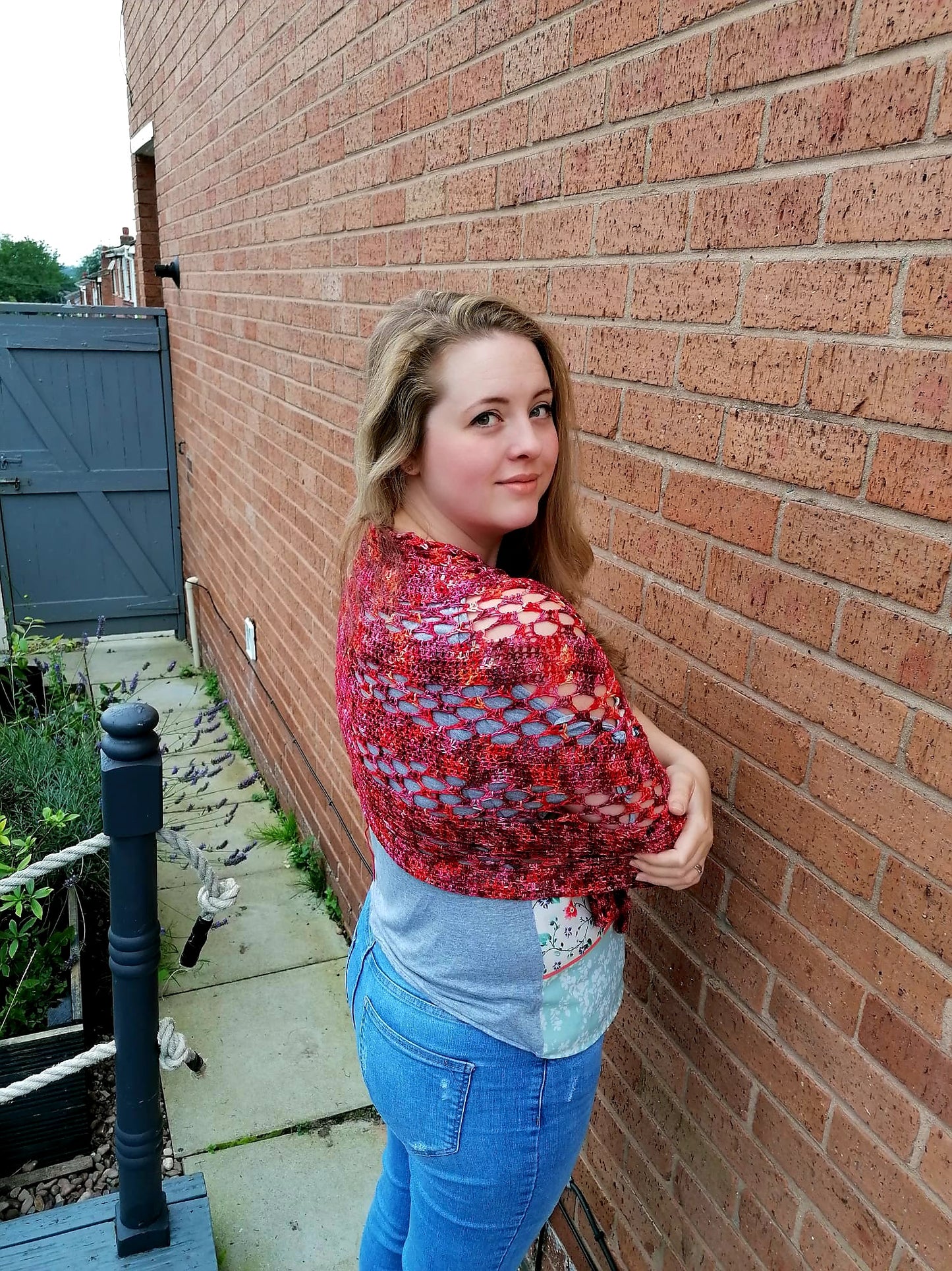 A woman is tanding against a wall wearing a red lace crochet wrap across her shoulders. She has her arms folded and is looking over her shoulder. The crochet shawl is wrapped tightly. 