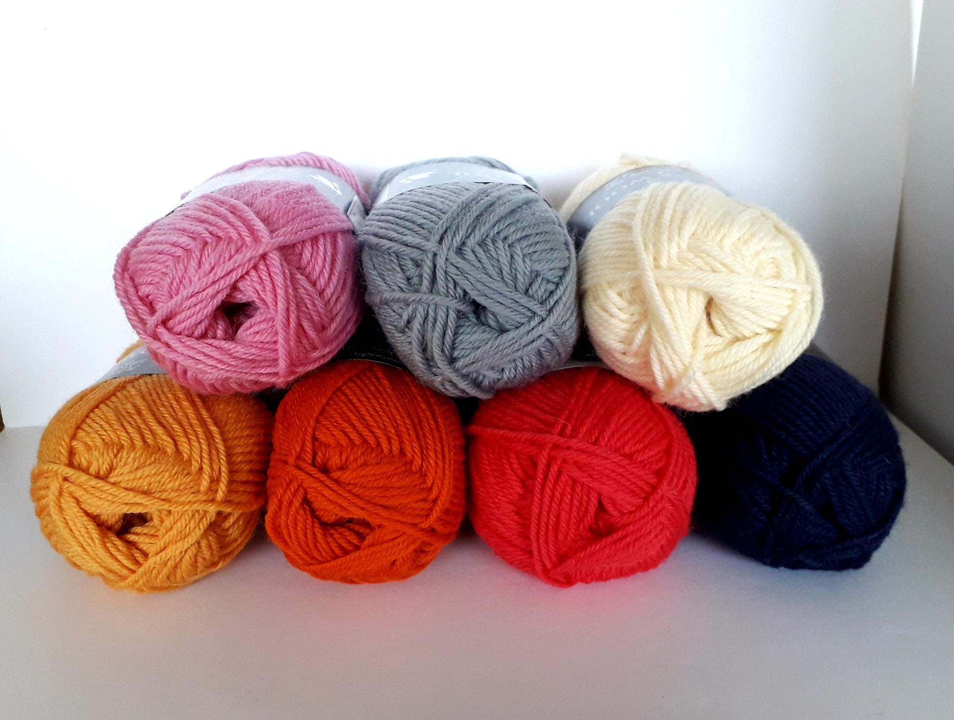 7 balls of yarn are stacked on top of each other. Their colours include white, grey, pink, orange, red, yellow and dark blue. 
