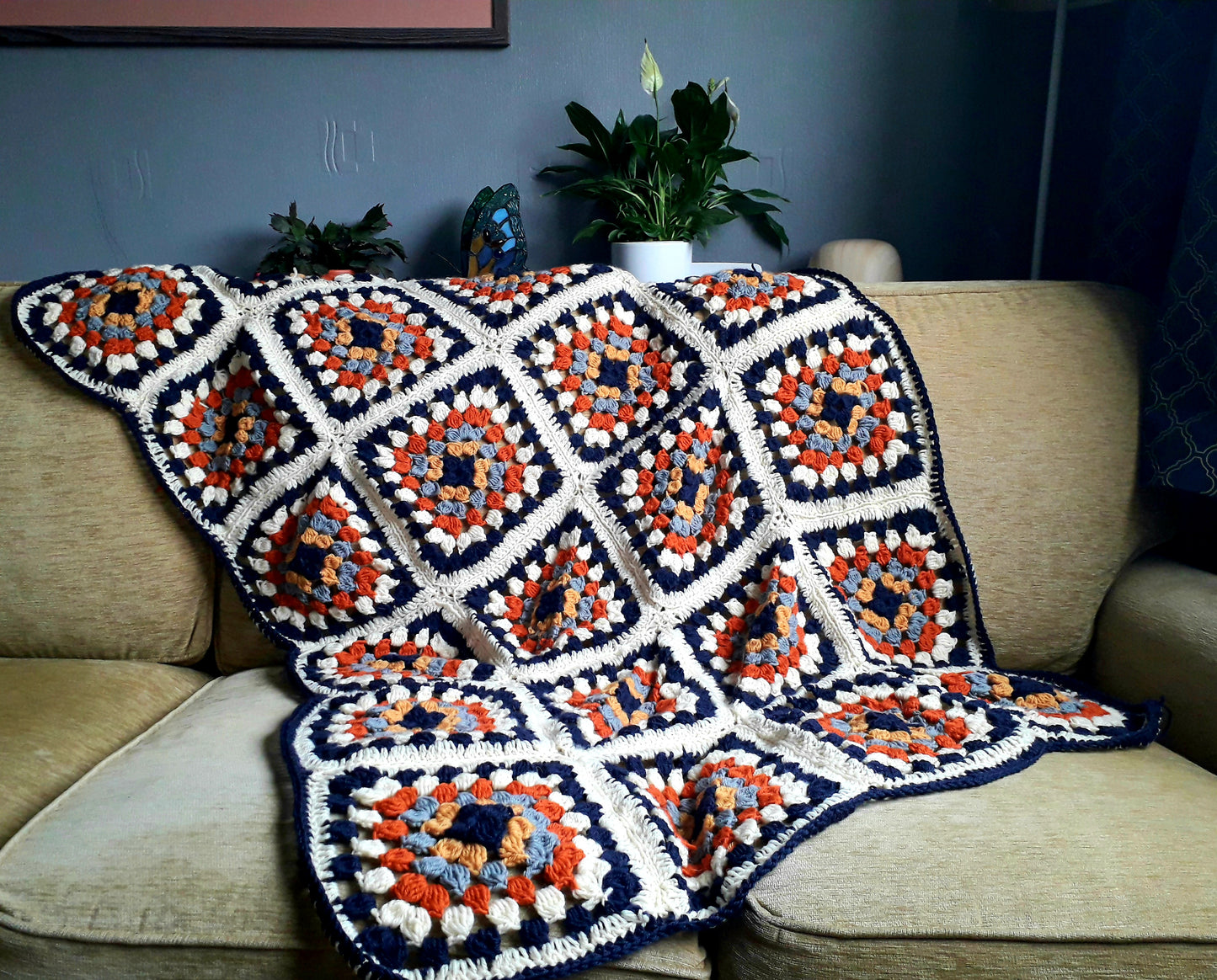 Image shows a granny square style blanket draped across a gold coloured sofa. The blanket has a navy blue border and is made up of 24 individual squares.